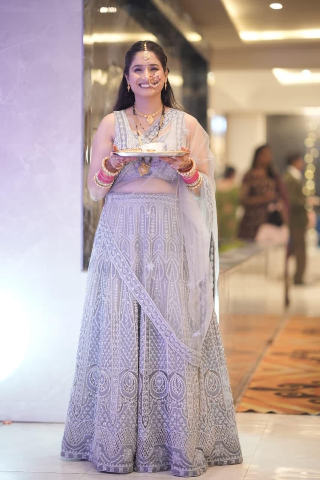 Full-length bridal look with soft glam makeup, silver-grey embroidered lehenga and gold jewellery, holding a puja thali