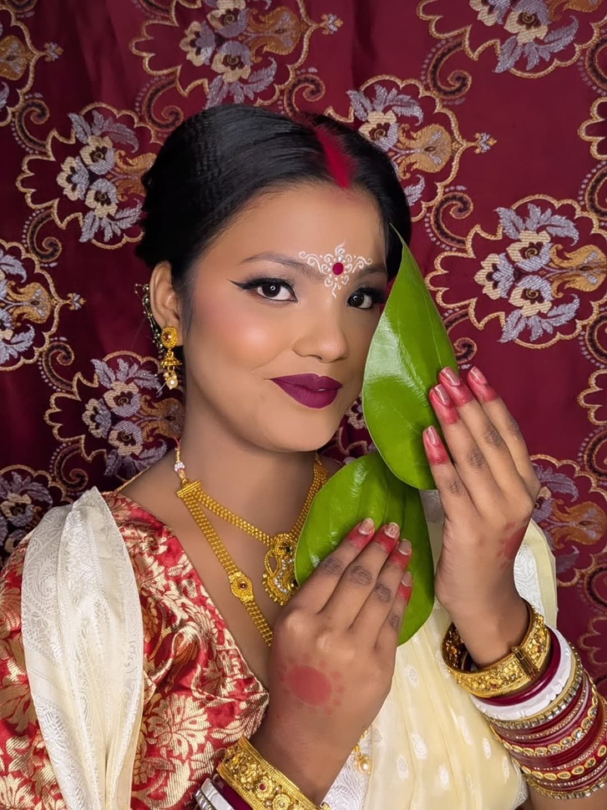 Bengali bridal makeup with bold plum lips, dramatic eyes, gold jewellery, and red sindoor on a bride in red and gold banarasi saree holding paan leaf