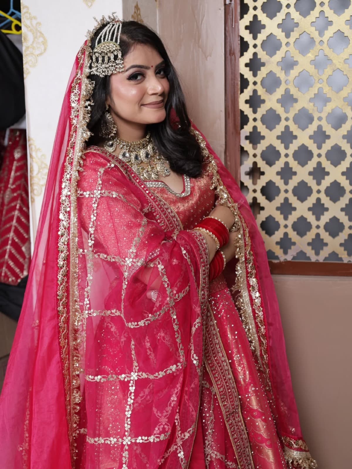 Full-length bridal shot with glowing makeup on a bride in a fuchsia pink lehenga with diamond-and-gold tiara and layered jewellery, ornate backdrop