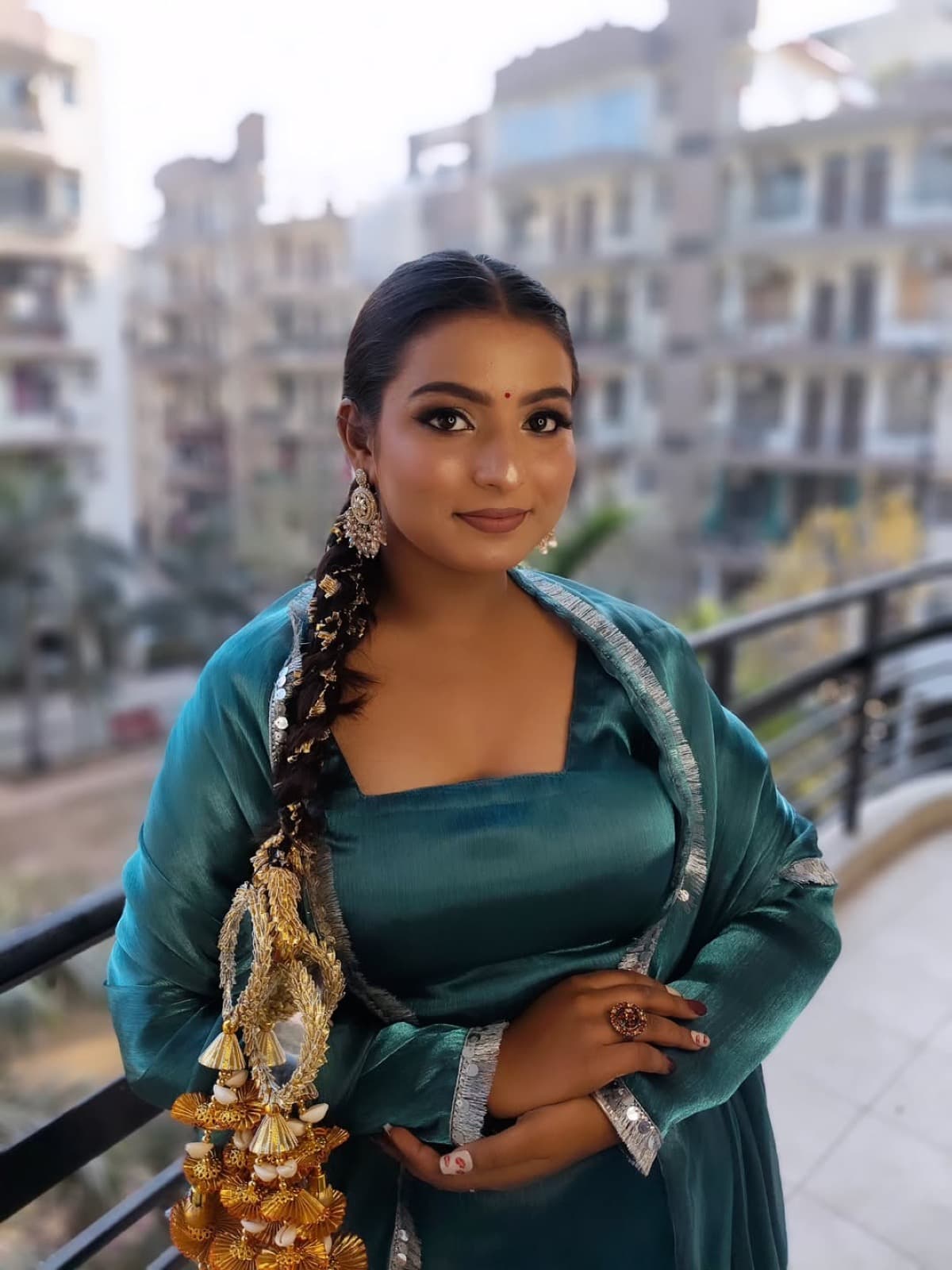 Festive makeup with warm eyes and glowing skin on a client in a teal silk lehenga with gold kaleere, standing on a balcony